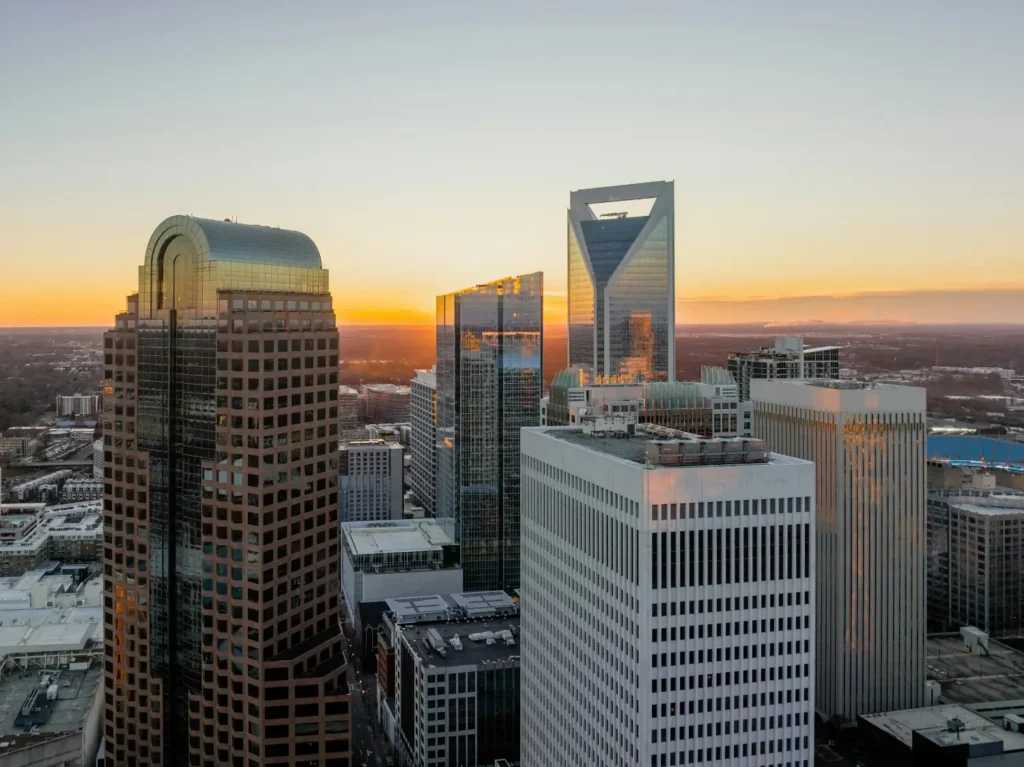Aerial view of downtown Charlotte, North Carolina skyline at sunset, featuring the city's distinctive modern skyscrapers including the Duke Energy Center with its curved glass facade and other prominent banking towers, all bathed in golden light against a gradient sky