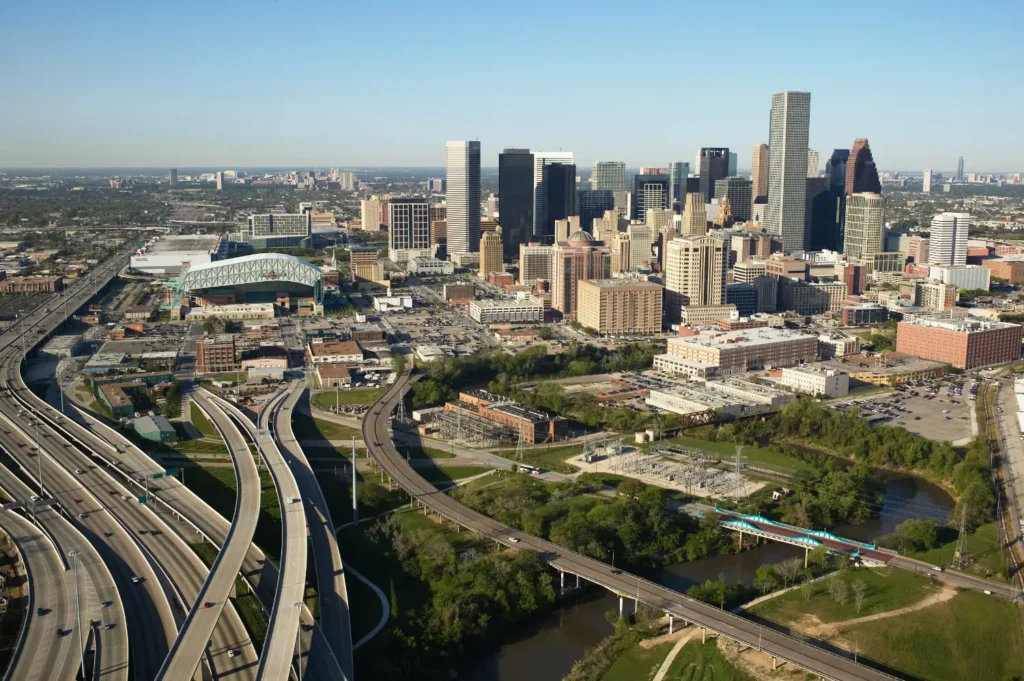 Aerial view of downtown Houston skyline with modern skyscrapers and office towers, featuring complex highway interchanges in the foreground, green parkland along a waterway, and the city's distinctive mix of glass and steel architecture under a clear blue sky