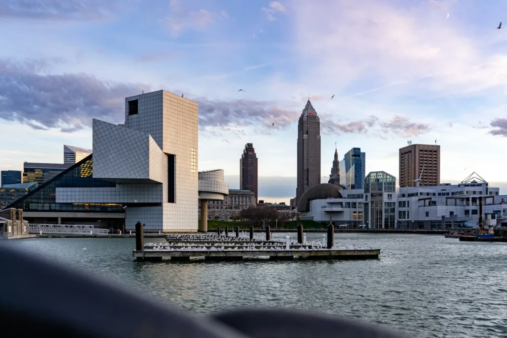 Cleveland skyline viewed from Lake Erie featuring the Rock and Roll Hall of Fame's distinctive pyramid-shaped glass building in the foreground, with downtown skyscrapers including Terminal Tower visible across the water under a dramatic cloudy sky at dusk